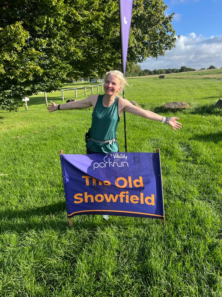 The Old Showfield parkrun, Woman stands by purple sign which reads The Old Showfield, parkrun, parkrun tourism, Running, Runner