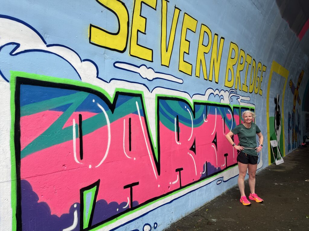 Severn Bridge parkrun, Severn Bridge parkrun B course, Parkrun, Parkrun tourism, A female runner in a green top black shorts and pink trainers stands by street art which says Severn Bridge parkrun
