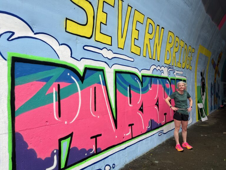Severn Bridge parkrun, Severn Bridge parkrun B course, Parkrun, Parkrun tourism, A female runner in a green top black shorts and pink trainers stands by street art which says Severn Bridge parkrun