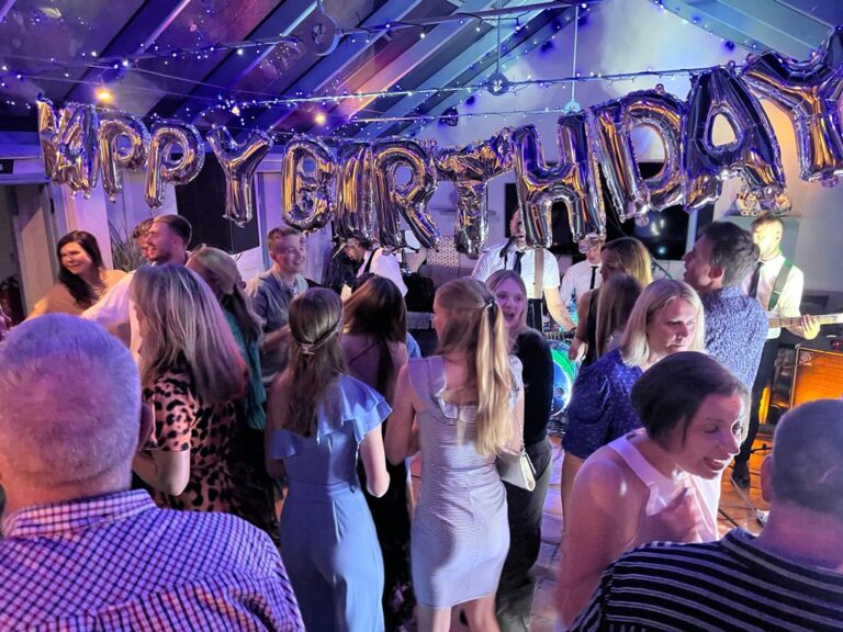 50th birthday party, Birthday party, 50th birthday, A group of people stand under silly Happy Birthday balloons and a band plays