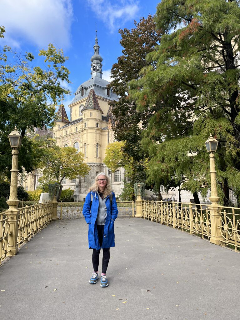 Vajdahunyad Castle, Budapest, Hungary, A woman in glasses and a blue coat stands on an ornate bridge outside a castle and surrounded by trees