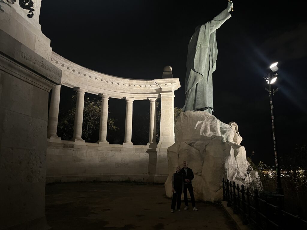 Gellert Monument, Budapest, Gellert, Statue, Things to do in Budapest, A man and a teenage girls stand next to a very large religious statue next to a series of pillars against a dark night sky