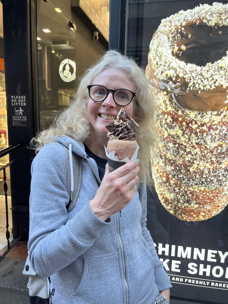 Chimney cake, Budapest, Hungary, A woman in a grey hoodie wearing glasses eats a chimney cake which looks like an ice cream cone filled with chocolate ice cream