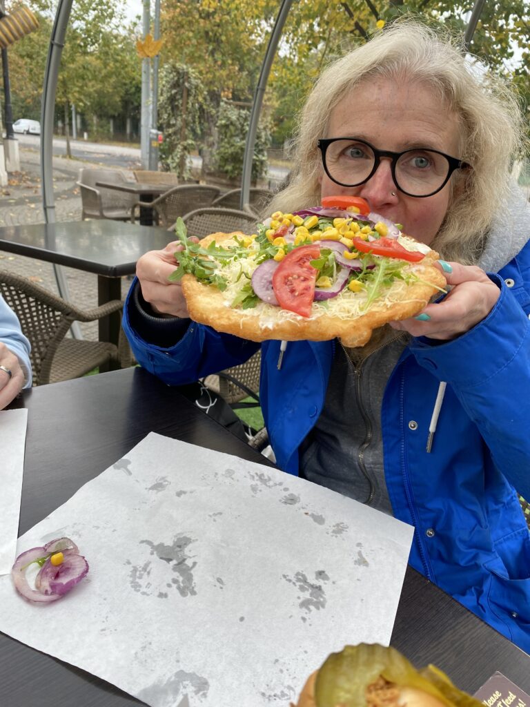 Langos, Budapest, A woman in a blue coat and glasses eats a langos covered in vegetables (a bit like a pizza)