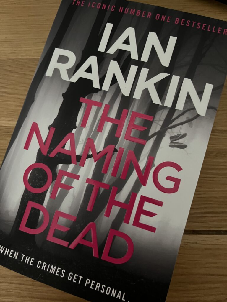 The Naming of the Dead by Ian Rankin, The Naming of the Dead, Ian Rankin, Book review, A book called The Naming of the Dead by Ian Rankin with a black and grey cover sits on a wooden table