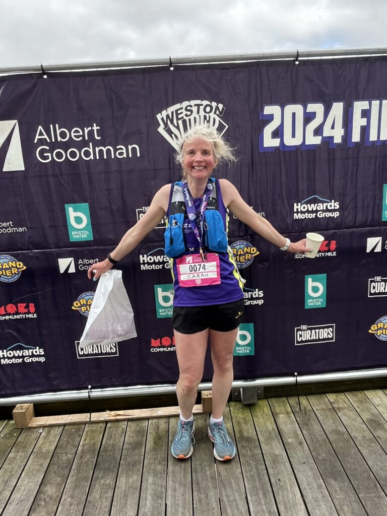 Weston Super Half finisher, Weston Super Half, Half marathon, Running, Runner, A smiling woman in shorts and a vest wearing a pink race number stands against a black backdrop covered in logos