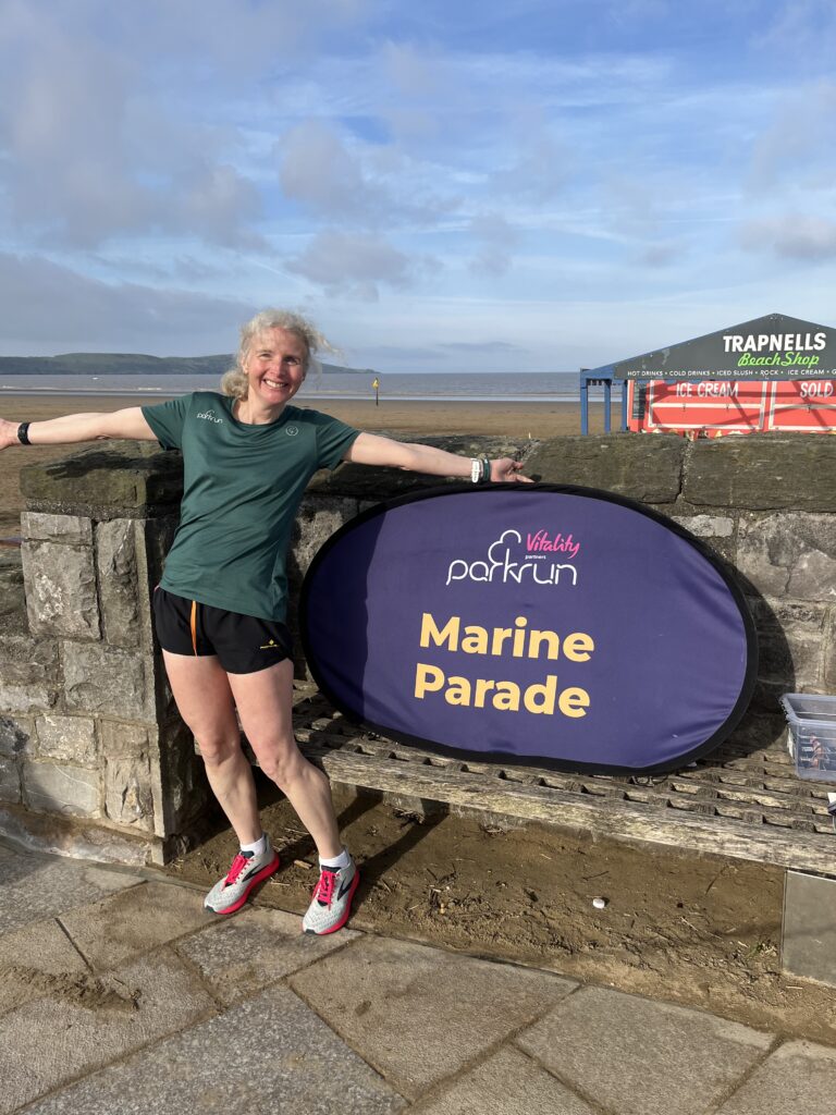 Marine Parade parkrun, Parkrun, Weston-super-Mare, Running, Runner, Parkrun tourism, Smiling woman in green Tshirt and black shorts stands by a big purple sign reading Marine Parade