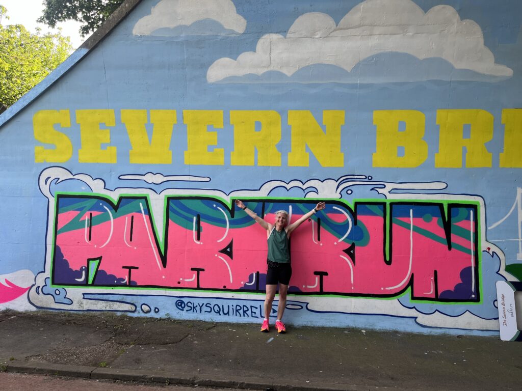 Severn Bridge parkrun, Runner, Running, Parkrun, Silent Sunday, My Sunday Photo, A female runner stands against a large mural reading Severn Bridge parkrun in colourful letters