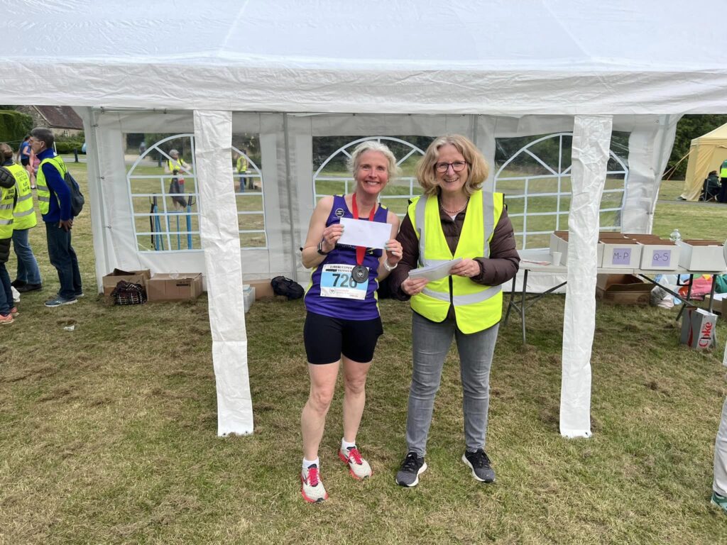 Cirencester 10k, Running, Runner, A smiling female runner holds up an envelope and stands net to a smiling woman in a high viz jacket