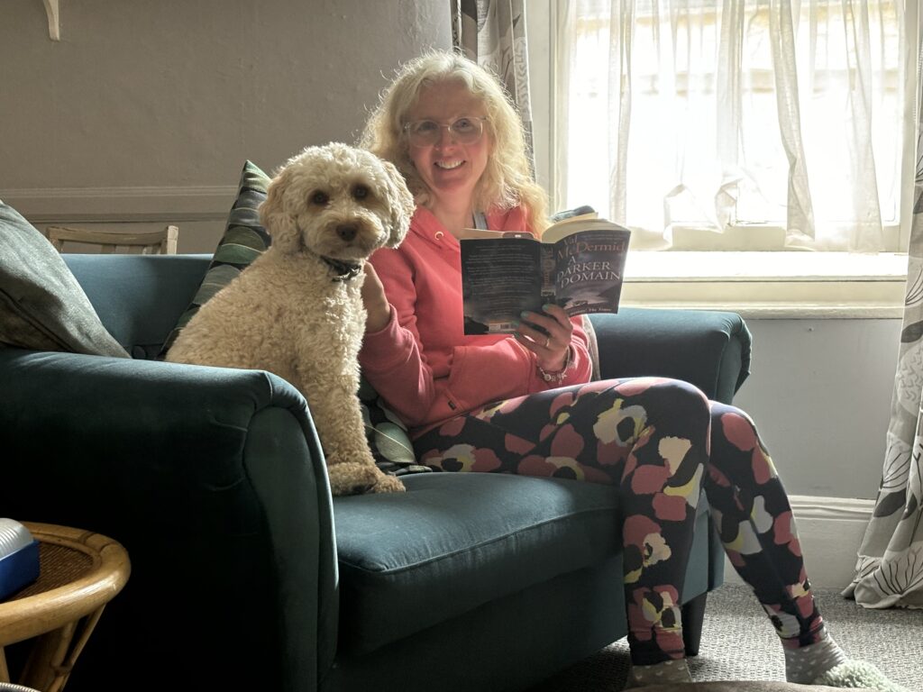 Airbnb, Hetty, Tenby, A smiling woman in a pink hoodie and colourful leggings sits on a sofa reading alongside a golden cockapoo dog