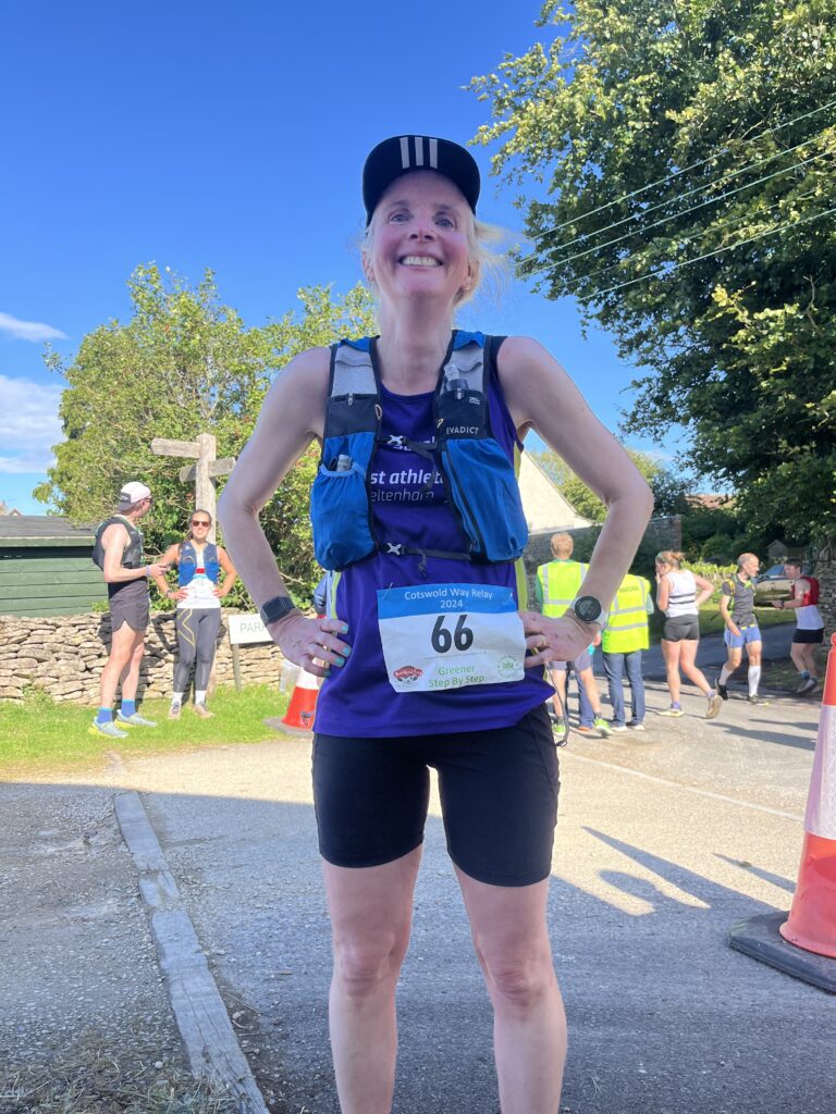 Cotswold Way Relay, Runner, Finish line, A smiling female runner in a black cap purple vest and black shorts stands on a country lane with other runners in the background