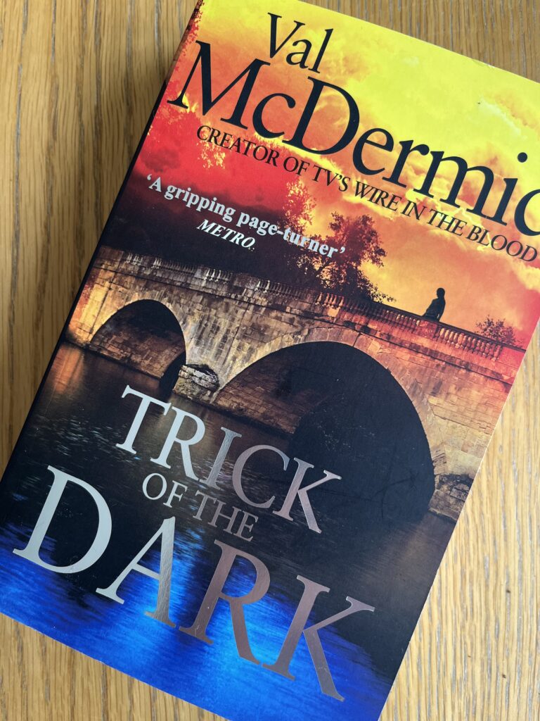 Trick of the Dark by Val McDermid, Trick of the Dark, Val McDermid Book review, A book with a bridge on the coloured cover called Trick of the Dark by Val McDermid sits on a wooden table