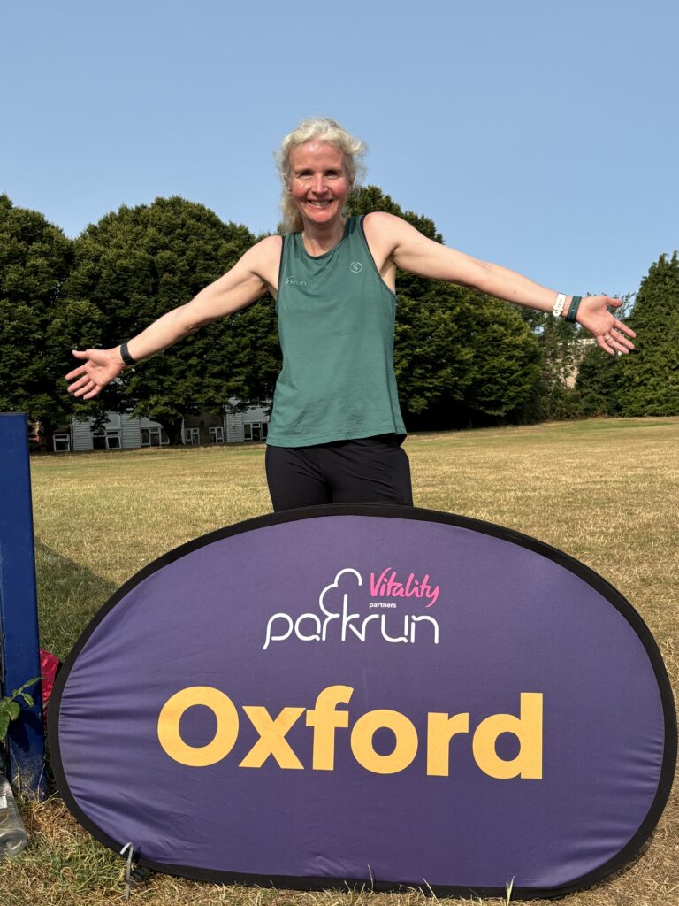 Oxford parkrun, Parkrun, Parkrun tourism, A smiling woman in a green vest stands behind a big purple sign which says Oxford