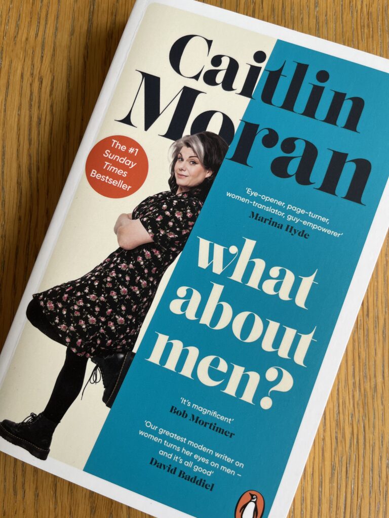 What About Men? by Caitlin Moran, What About Men?, Caitlin Moran, Book review, A book called What About Men? by Caitlin Moran with a photo of a middle aged woman leaning on the front cover sits on a wooden table