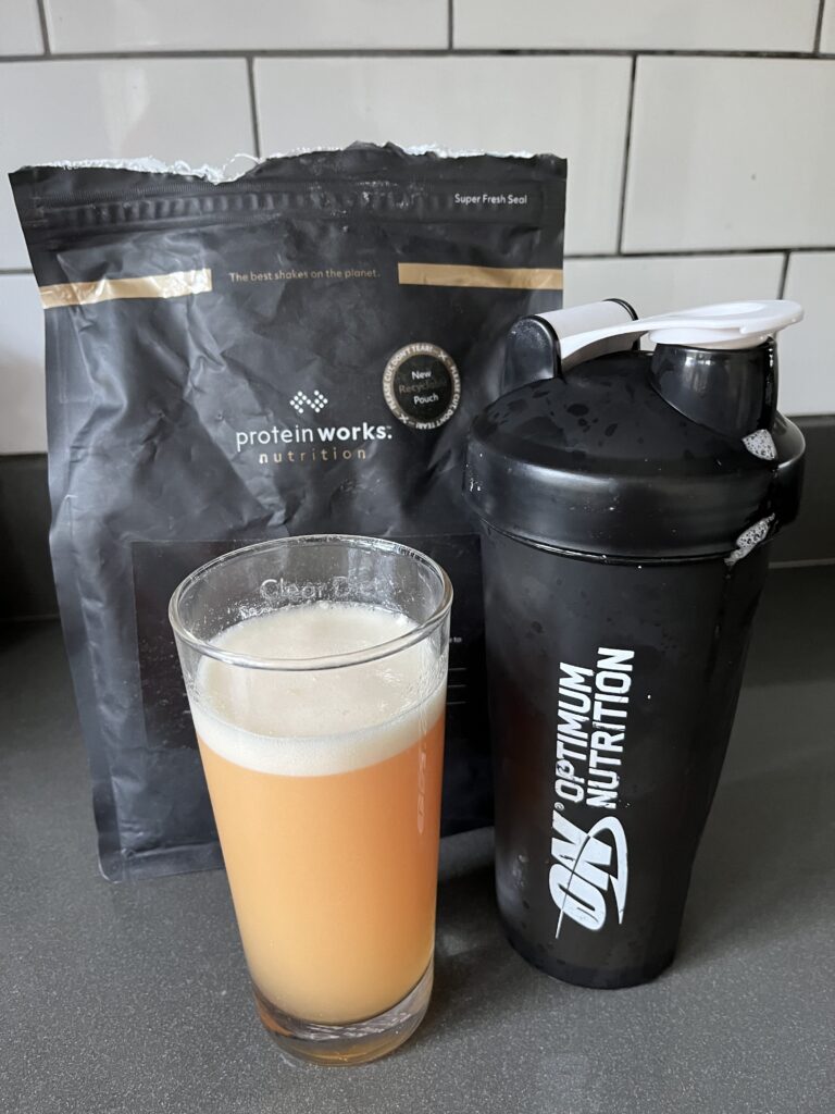 Protein shake, Clear protein, Protein works clear protein, A glass of orange froth liquid sits next to a black plastic cup with a black lid and in front of a black packet which says protein works on the front