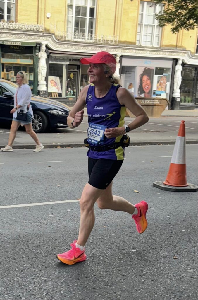 Cheltenham half marathon 2024, Running, Runner, A smiling female runner in a purple vest and pink hat and trainers runs along a road past some shops
