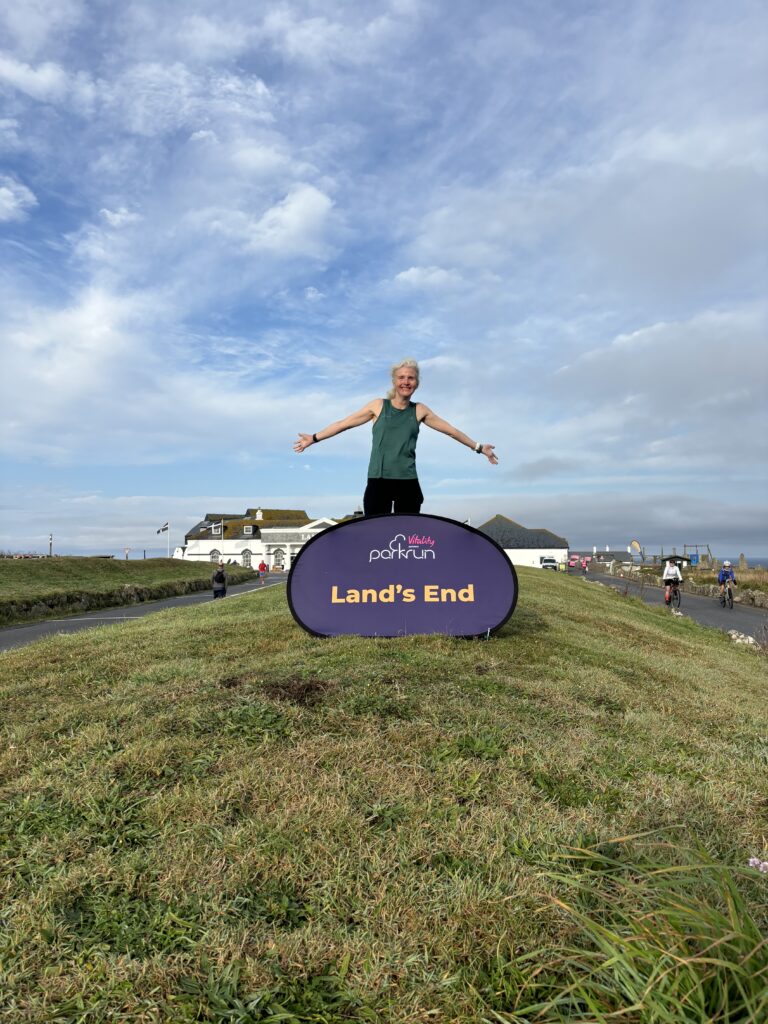 Land's End parkrun, Land's End, Cornwall, Parkrun, Parkrun tourism, A smiling female runner in a green vest stands behind a big purple sign which says Land's End there is a big blue sky and some white buildings in the background