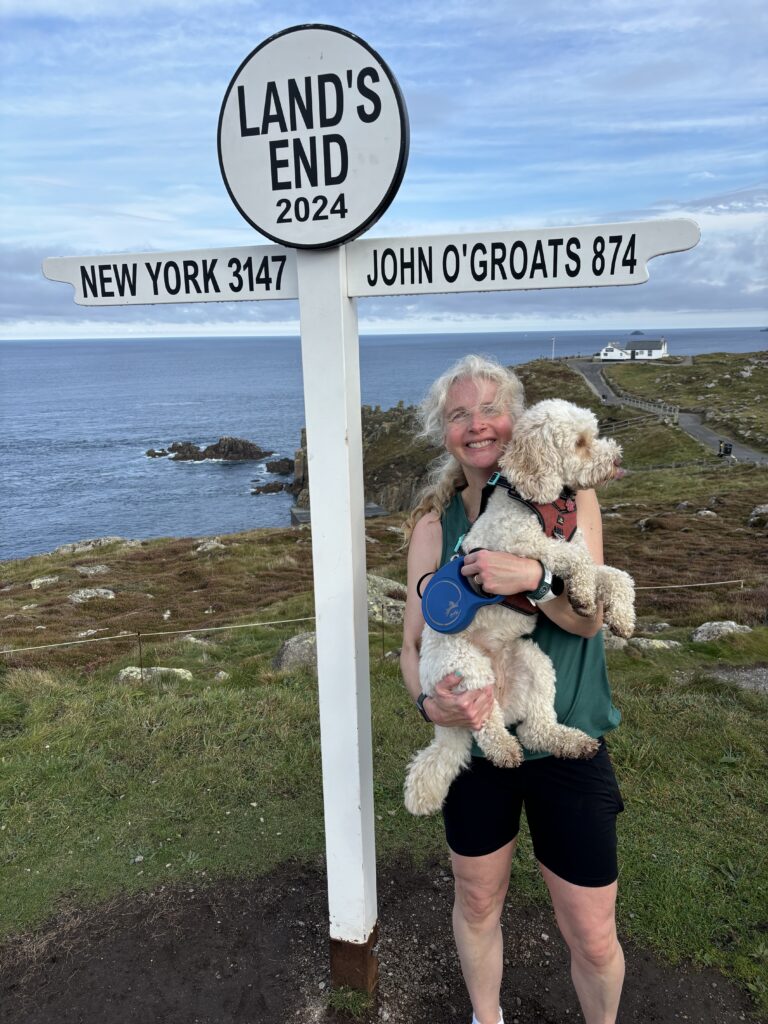 Land's End parkrun, Land's End, Cornwall, Parkrun, Parkrun tourism, Land's End sign, Cockapoo, A smiling woman in shorts and a green vest holds a golden cockapoo dog next to the famous Land's End sign with rocks and sea in the background