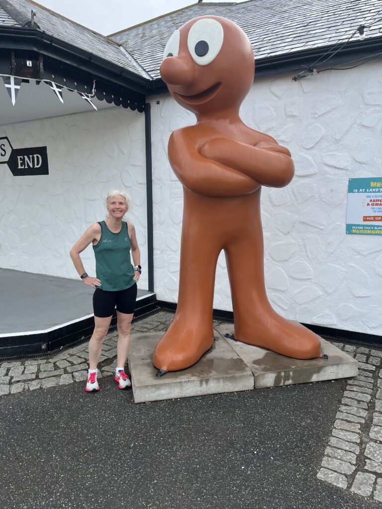 Land's End parkrun, Land's End, Cornwall, Parkrun, Parkrun tourism, Morph, Aardman, A smiling woman in shorts and a green vest stands next to a very tall orange statue of a cartoon man with his arms folded
