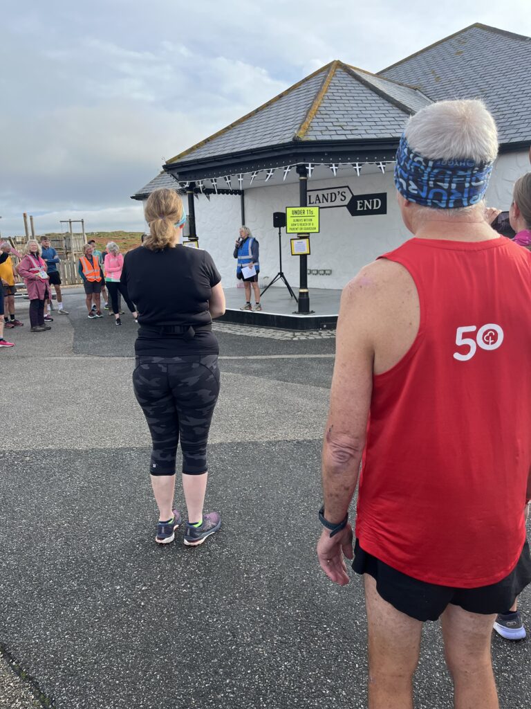 Land's End parkrun, Land's End, Cornwall, Parkrun, Parkrun tourism, Rear view of a group of runner withe a bandstand in the background and a woman in a blue hi-vis vest talking to them