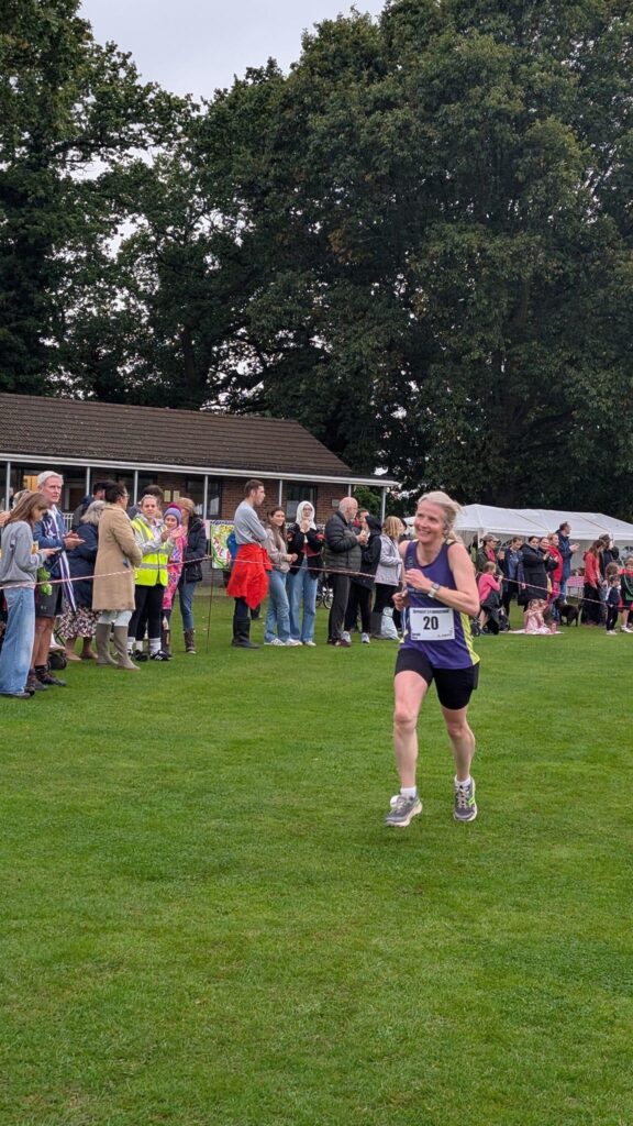 Apperley Quarter Marathon, Running, Runner, Apperley, Gloucestershire, A smiling female runner in a purple vest runs along the grass in front of crowds of people
