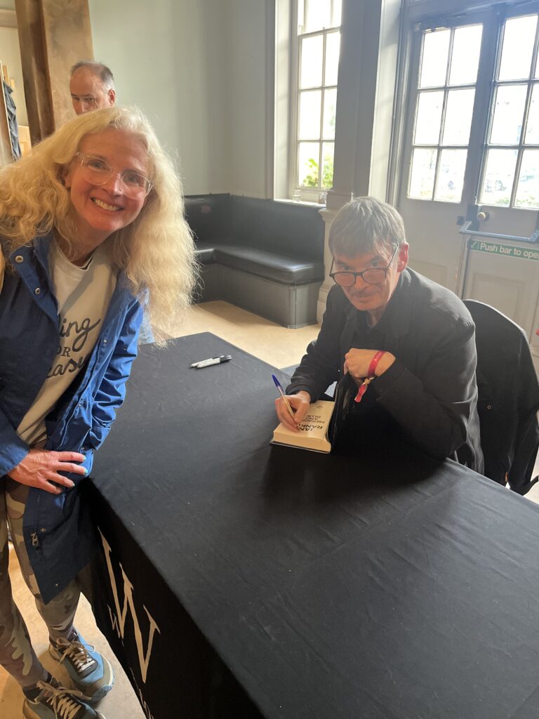 Midnight and Blue, Ian Rankin, Cheltenham Literature Festival, A smiling blonde woman in glasses wearing a blue coat stands next to a table covered in a black cloth where a man dressed in black signs a book