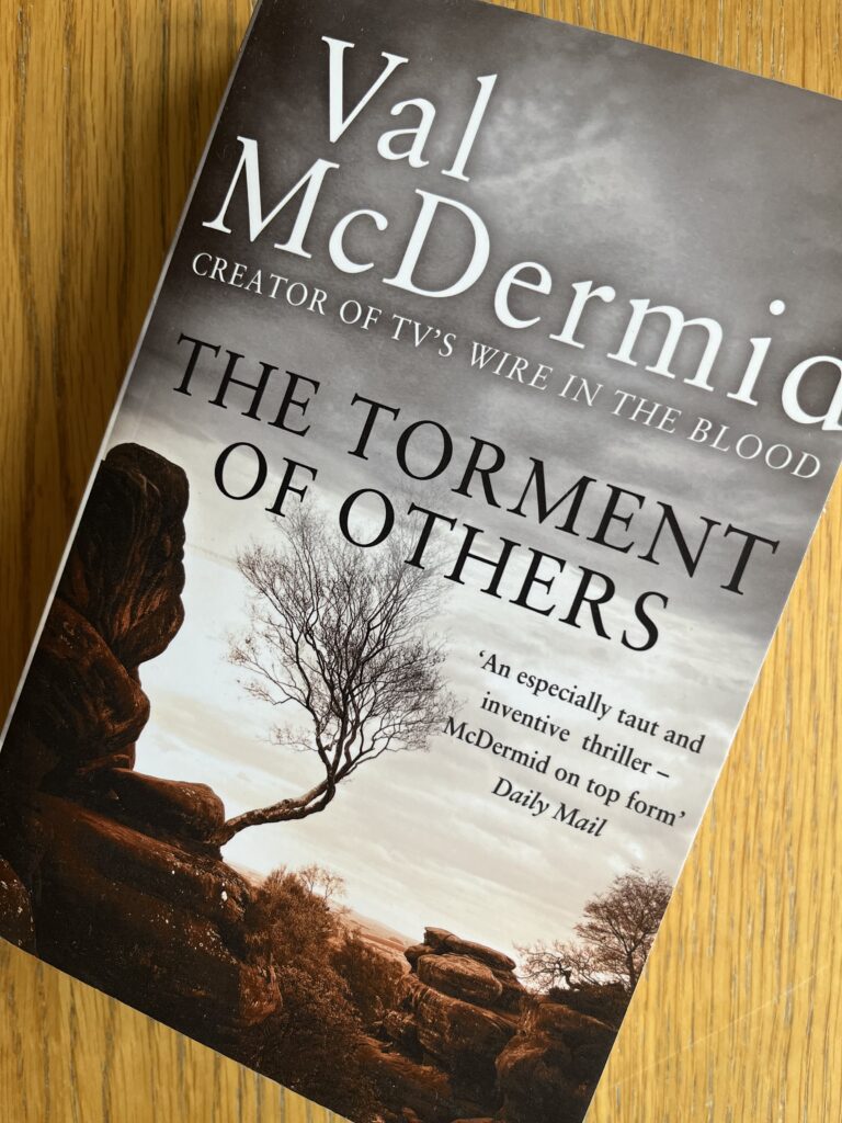 The Torment of Others by Val McDermid, The Torment of Others, Val McDermid, Book review, A grey book with a bare tree on the cover called The Torment of Others by Val McDermid sits on a wooden table
