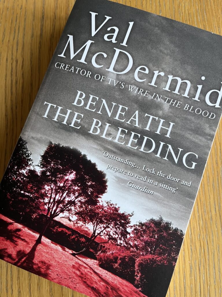 Beneath the Bleeding by Val McDermid, Beneath the Bleeding, Val McDermid, Book review, A grey and red book called Beneath the Bleeding by Val McDermid sits on a wooden table