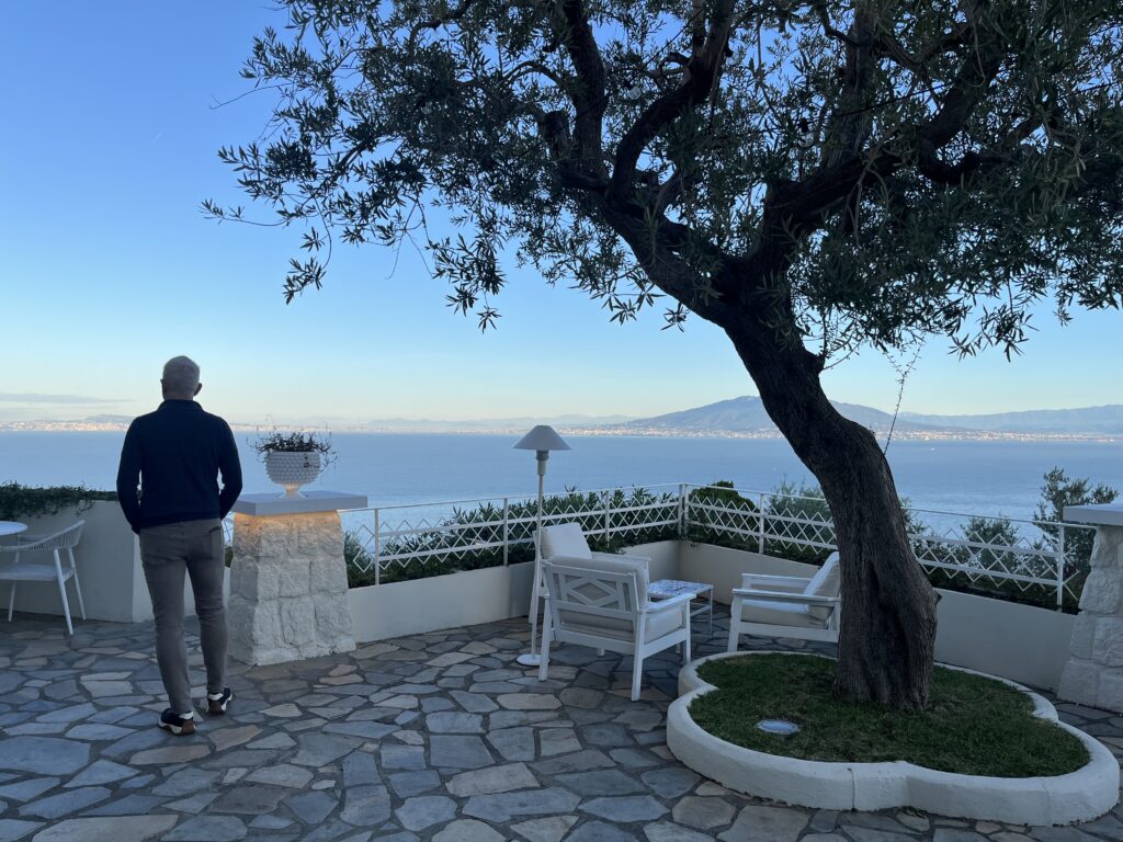 Sorrento, Sunrise, Grand Hotel Aminta, Italy, A man stands on a terrace with tables chairs and an olive tree and looks out to sea