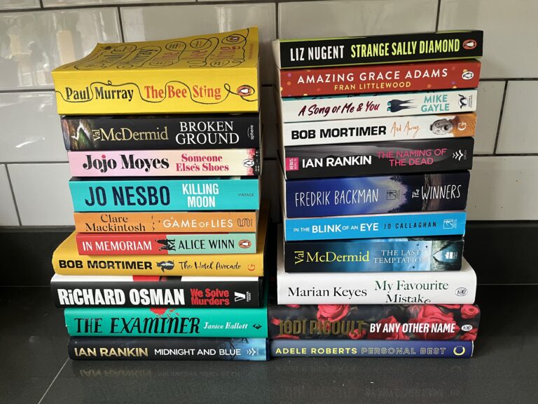 Books I read in 2024, Books, Reading, Two piles of books with covers in a variety of colours sit on a dark surface with white tiles in the background
