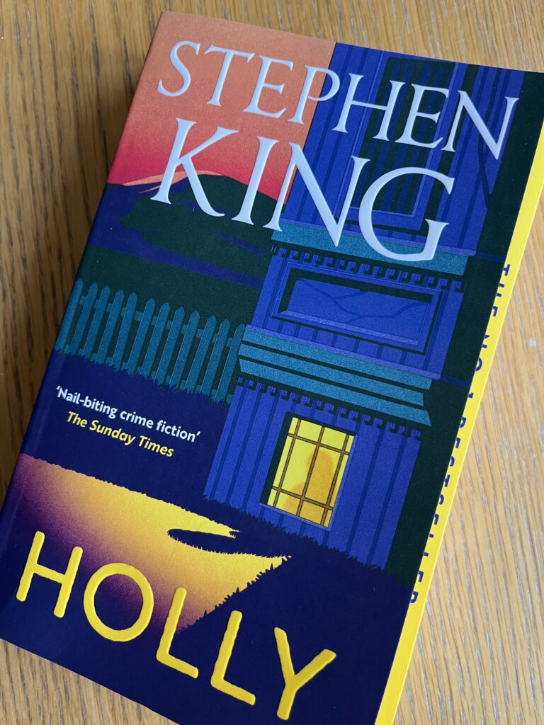 Holly by Stephen King, Holly, Stephen King, Book review, A book called Holly by Stephen King with a mainly blue cover featuring a graphic image of a house with a lit window sits on a wooden table
