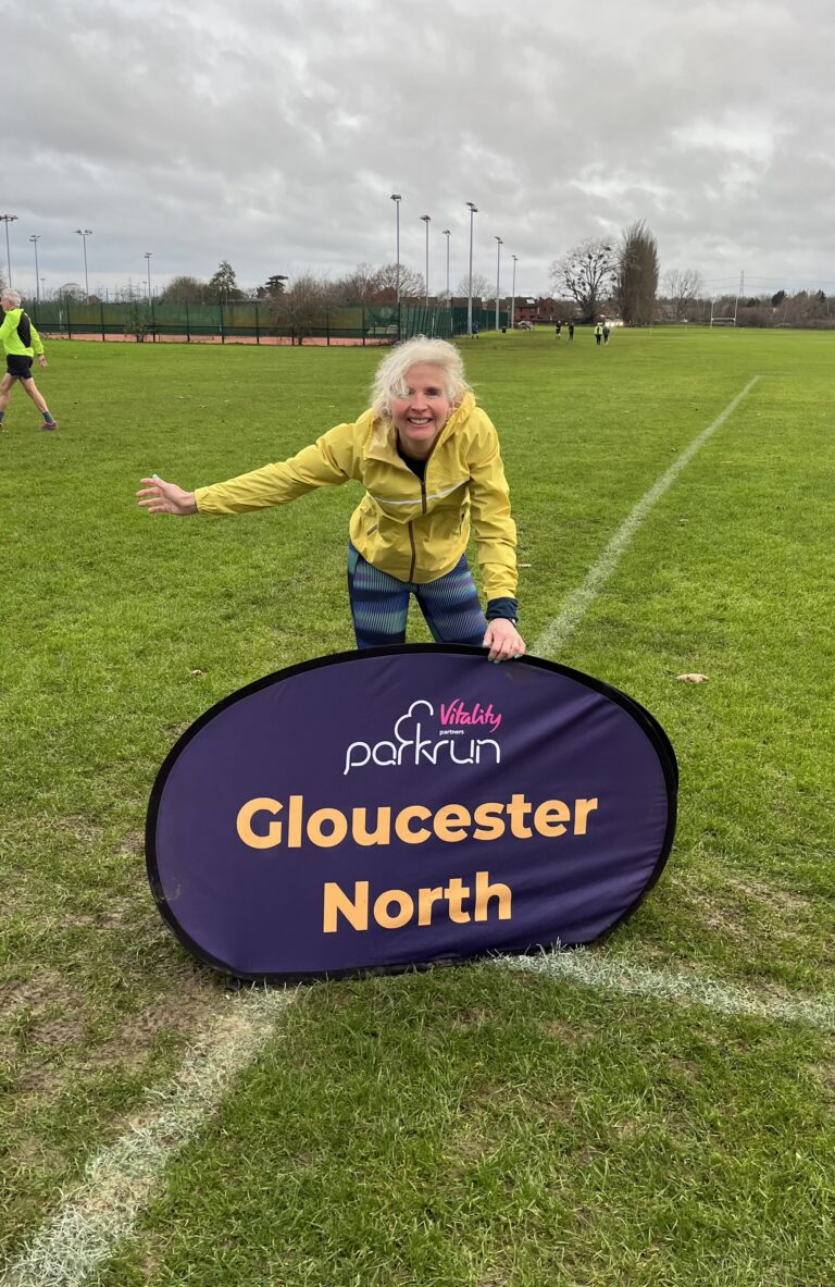 Gloucester North parkrun, Parkrun tourism, Parkrun, Running, Runner, A smiling female runner in a yellow coat stands on grass holding on to a big purple sign which says Gloucester North