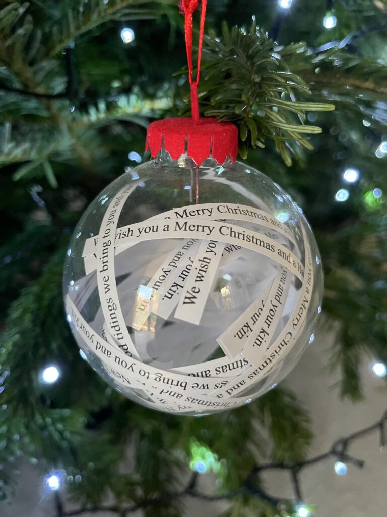 Christmas tree, Christmas bauble, Christmas decorations, Silent Sunday, My Sunday Photo, A clear glass Christmas tree with a red ribbon and lots of strips of paper inside it saying We wish you a Merry Christmas hangs from a real Christmas tree with white lights in the background