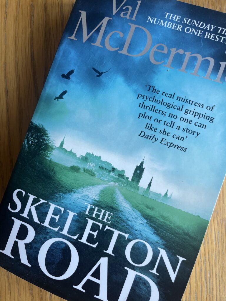 The Skeleton Road by Val McDermid, The Skeleton Road, Val McDermid, Book review, A blue book with a silhouette of a city on the cover called The Skeleton Road by Val McDermid sits on a wooden table