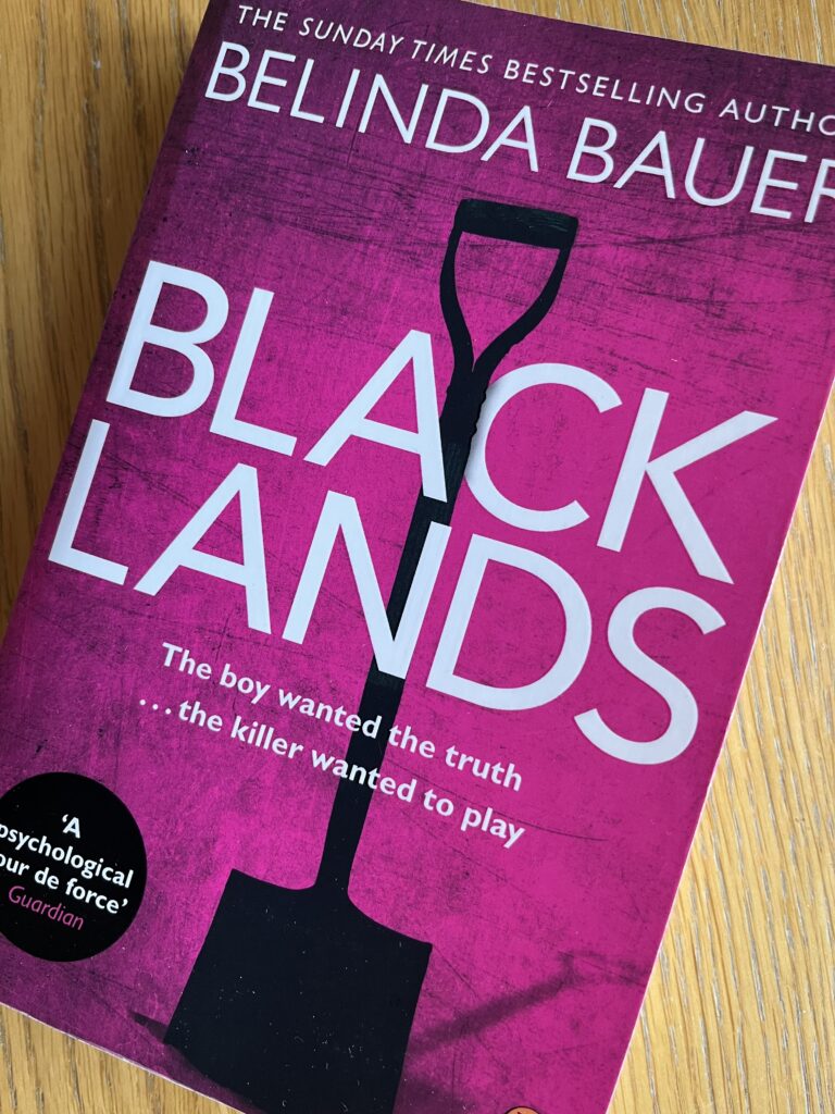 Blacklands by Belinda Bauer, Blacklands, Belinda Bauer, Book review, A pink book with a silhouette of a spade on the cover called Blacklands by Belinda Bauer sits on a wooden table