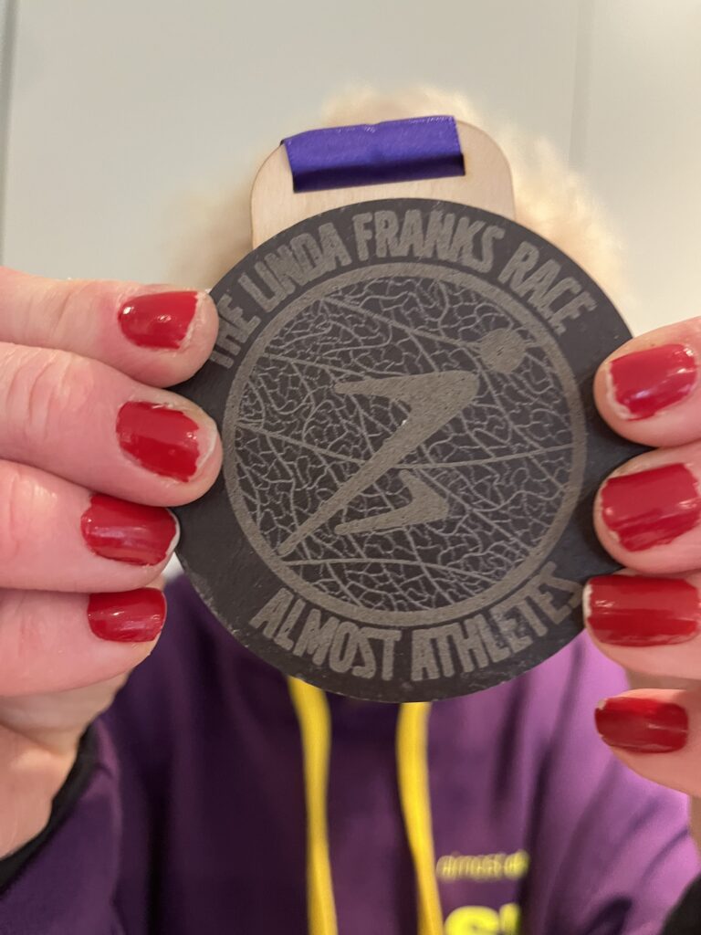 The Linda Franks race, Medal, Running, Runner, Silent Sunday, My Sunday Photo, Close up of a dark grey medal with a purple ribbon which says Linda Franks Race held by a woman with red nails