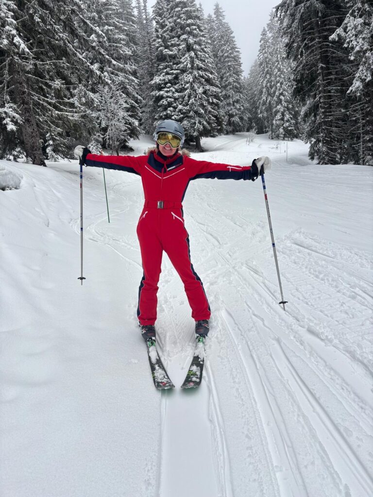 Skiing, Skiing holiday, A female skier in a red suit holds her poles out wide and stands on the snow surrounded by snow covered trees