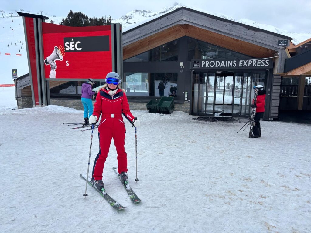 Skiing, Skiing holiday, Avoriaz, A female skier in a red suit wearing a helmet and blue goggles stands on the snow in front of a building which says Prodains Express