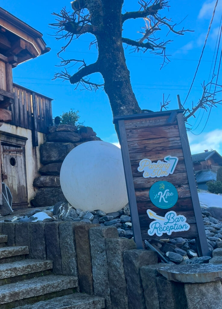Hotel, Hotel Mazots des Fees, Morzine, A wooden sign which says Bar and Reception in front of a bare tree at the bottom of some stone steps with a wooden building and blue sky in the background
