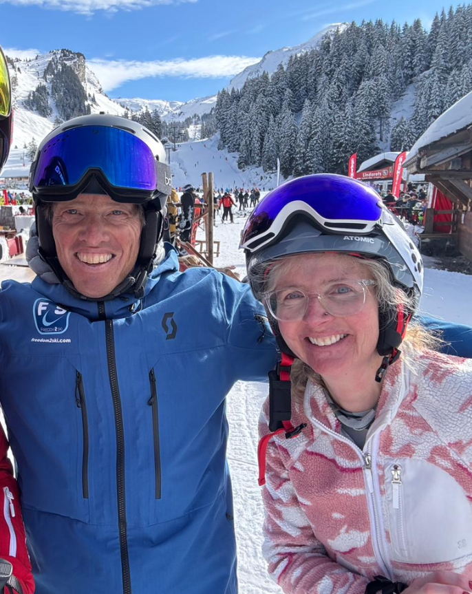 Skiing, Skiing holiday, Ski instructor, A man in a blue coat with a helmet and blue goggles on his head stands with a woman in a pink and white fleece wearing a helmet and blue goggles on her head, they are surrounded by snow, mountains and snow covered trees