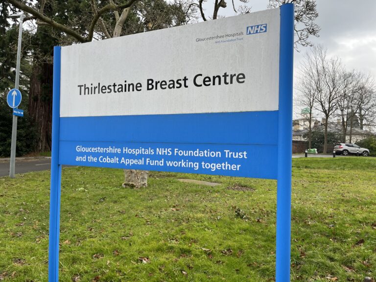 Thirlestaine Breast Centre, NHS breast screening, Mammogram, Gloucestershire, A large blue and white sign standing on grass with trees in the background and the sign says Gloucestershire Hospitals NHS Trust Thirlestaine Breast Centre