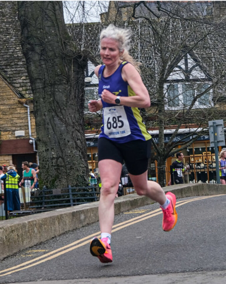 Bourton 10k, Running, Runner, A female runner in a purple vest and black shorts runs down a road with buildings and bare trees in the background