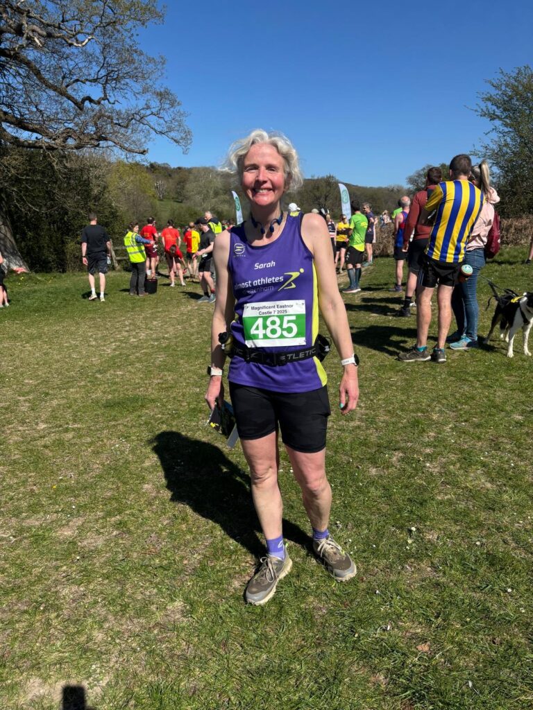 Running, Magnificent Eastnor 7, Runner, A smiling female runner in a purple vest and black shorts stands on grass surrounded by trees and blue sky with other runners in the background