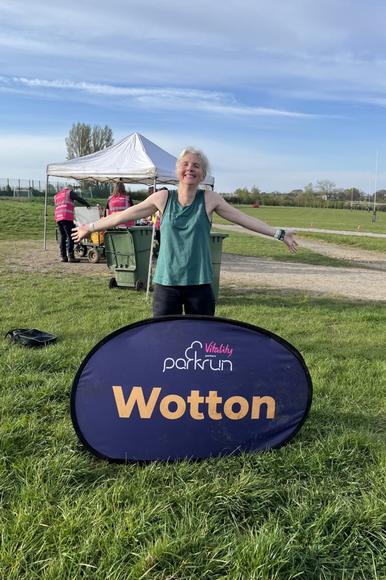 Wotton parkrun, Parkrun tourism, Running, Runner, A smiling female runner in a green vest stands on grass behind a big purple sign which says Wotton in yellow