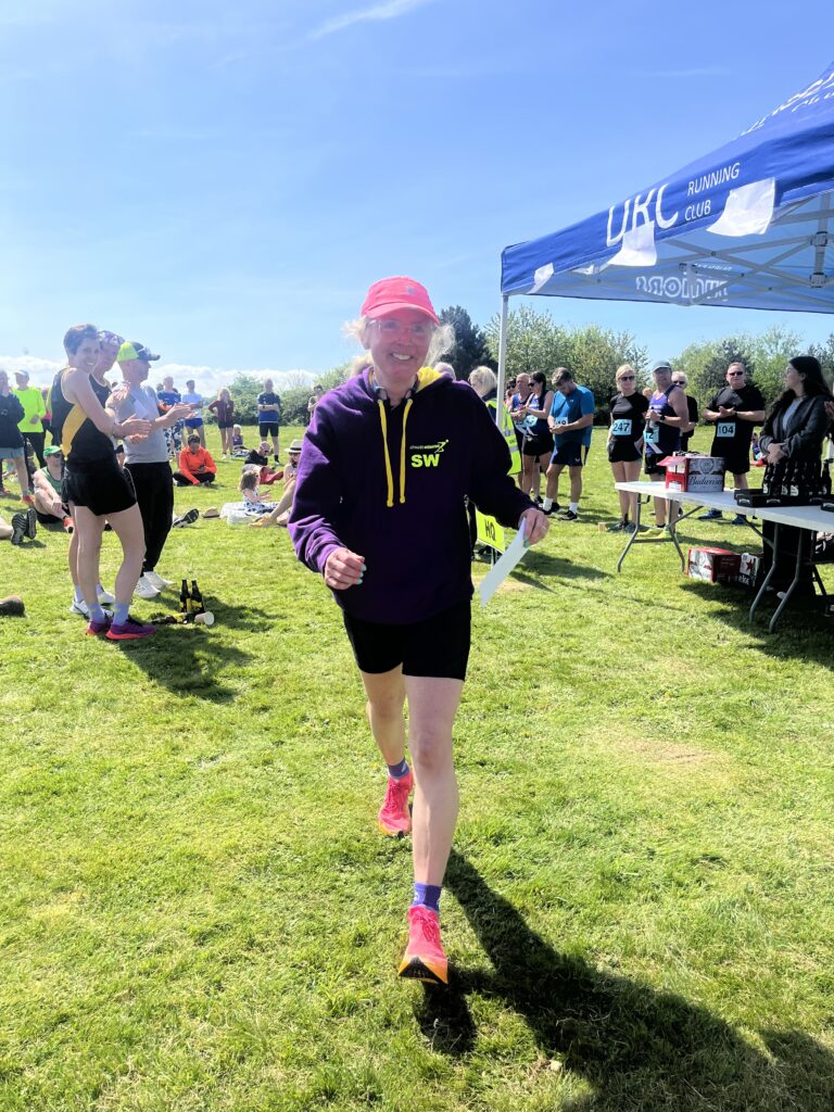 Berkeley 10k, Berkeley 10k prize, A smiling woman in black shorts and a purple hoodie holds a white envelope with lots of other runners in the background