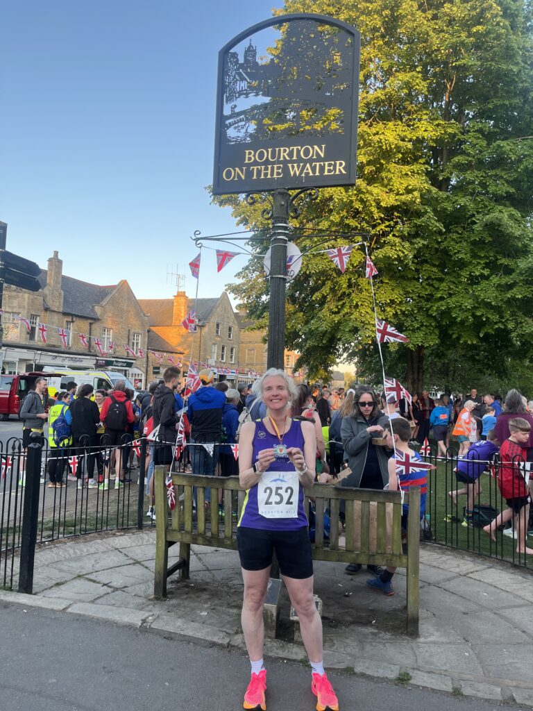 Bourton mile, Running, Runner, Bourton-on-the-Water, Gloucestershire, Cotswolds, A smiling female runner in a purprle vest and black shorts wearing a medal stands in front of a metal sign which says Bourton on the Water in front of crowds of people