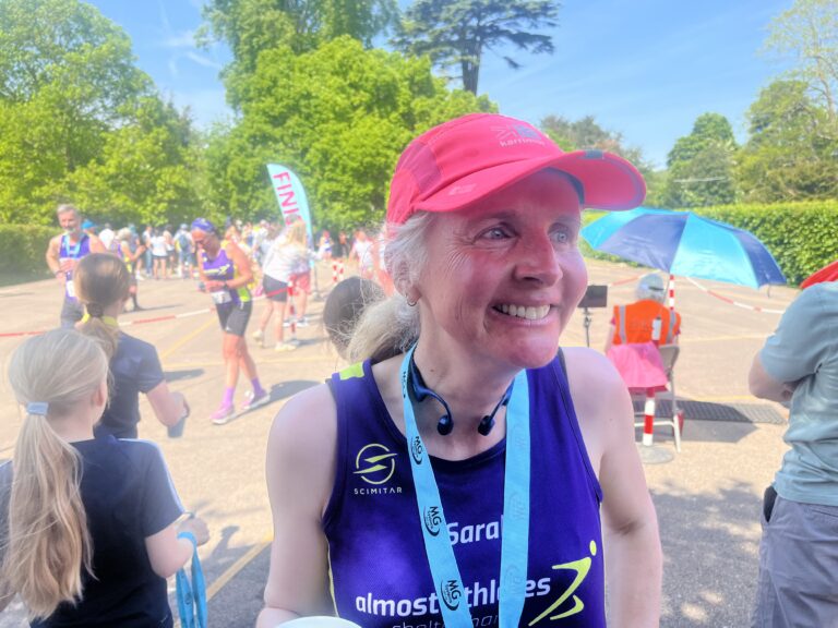 Sherborne 5 miler, Running, Race, Head and shoulders image of a smiling female runner in a purple vest and ink hat with crows of people and a finish line visible in the background