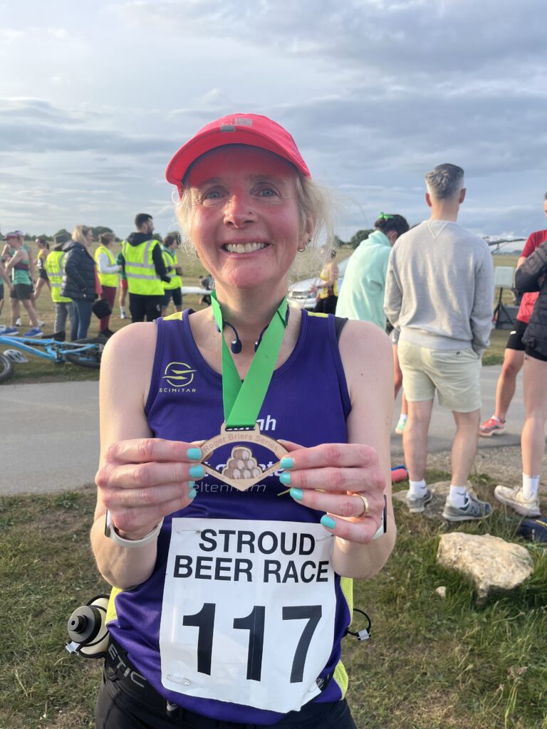 Stroud Beer Race, Medal, Runner, Running, A smiling female runner in a pink cap and purple vest wearing a number holds up a small wooden medal