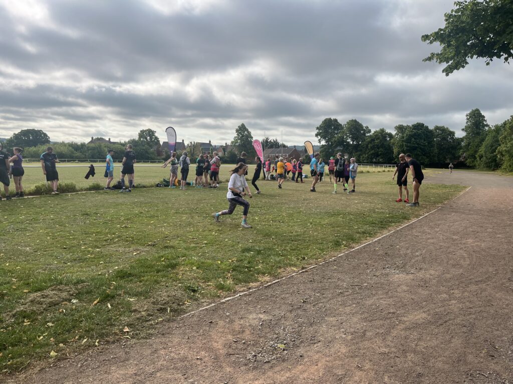 King George V Playing Field parkrun, Parkrun, Parkrun tourism, Runners, A group of runners standing around on grass surrounded by trees with three flags along the grass