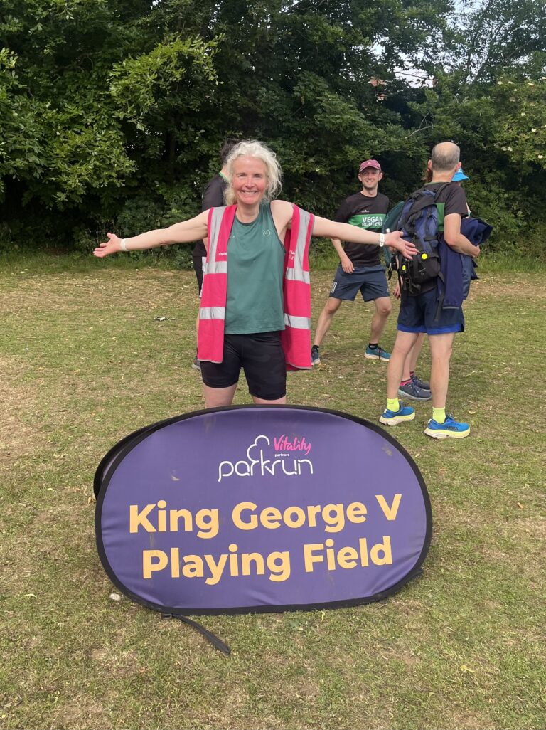 King George V Playing Field parkrun, Parkrun, Parkrun tourism, Cheltenham, Running, Runner, A smiling woman in pink hi viz over a green vest and black shorts stands behind a purple sign which says King George V Playing Field